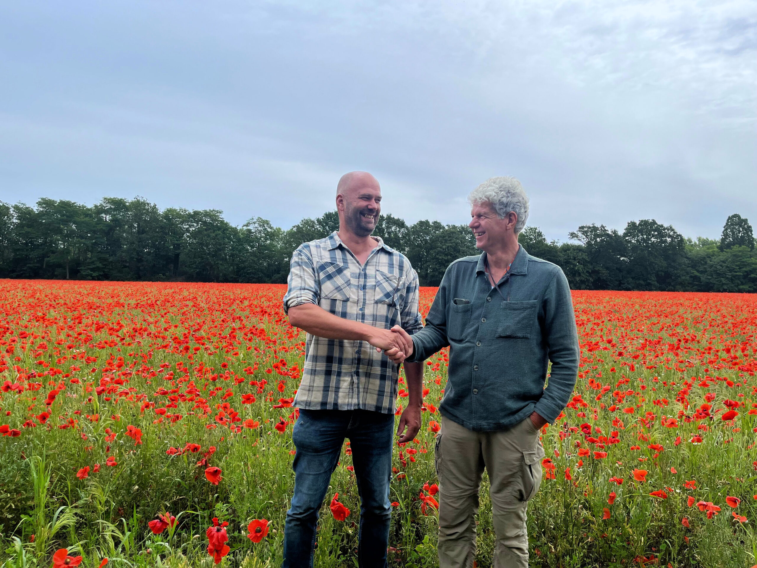 Egbert Hogendoorn en Peter de Groot schudden elkaar lachend de hand in een veld met bloeiende klaprozen.